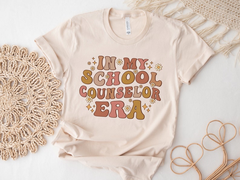 May include: A beige t-shirt with the text "In My School Counselor Era" in a retro font with pink, orange, and yellow colors. The text is surrounded by small white flowers and gold glitter.