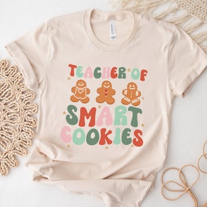 May include: A beige t-shirt with the text "Teacher of Smart Cookies" in colorful, retro-style lettering. Three gingerbread men are depicted above the text.