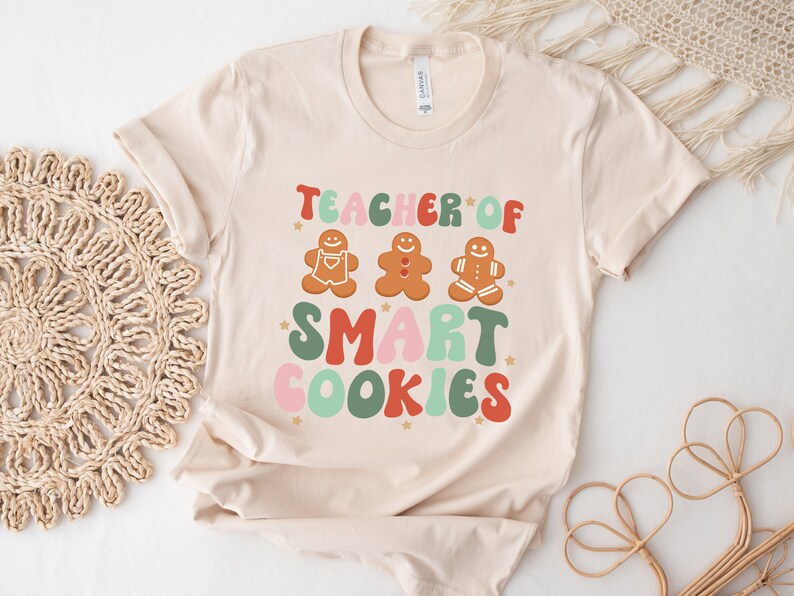 May include: A beige t-shirt with the text "Teacher of Smart Cookies" in colorful, retro-style lettering. Three gingerbread men are depicted above the text.