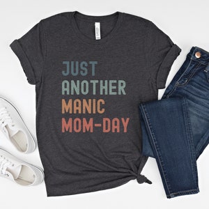 May include: A dark grey t-shirt with the text "JUST ANOTHER MANIC MOM-DAY" in a rainbow colour scheme. The shirt is paired with blue jeans and white trainers.