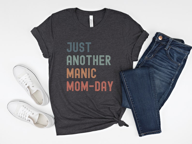 May include: A dark grey t-shirt with the text "JUST ANOTHER MANIC MOM-DAY" in a rainbow colour scheme. The shirt is paired with blue jeans and white trainers.