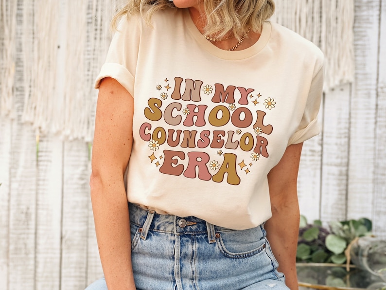May include: A beige t-shirt with the text "In My School Counselor Era" in a retro font with pink, brown, and gold accents. The text is surrounded by small white flowers and gold stars.
