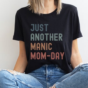 May include: A black t-shirt with the text "JUST ANOTHER MANIC MOM-DAY" in a vintage style font with a faded colour effect.