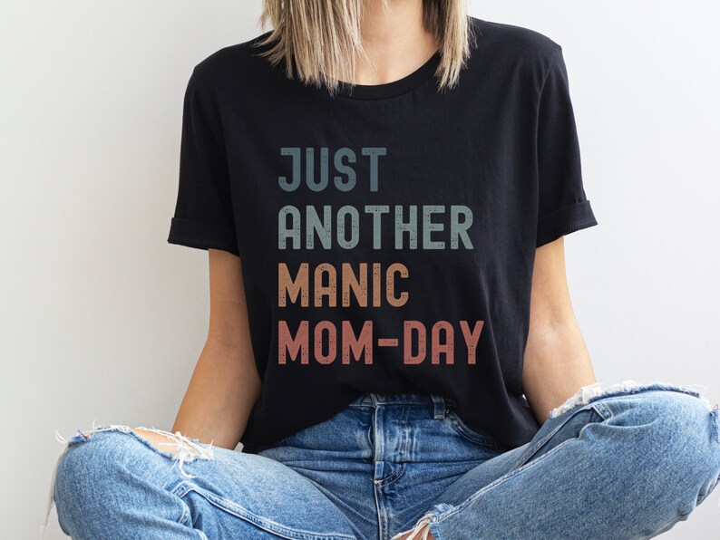May include: A black t-shirt with the text "JUST ANOTHER MANIC MOM-DAY" in a vintage style font with a faded colour effect.
