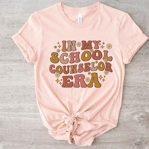 May include: A pink t-shirt with the text "In My School Counselor Era" in a retro font with brown, orange, and yellow colors. The text is surrounded by small white flowers and stars.