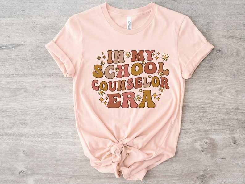 May include: A pink t-shirt with the text "In My School Counselor Era" in a retro font with brown, orange, and yellow colors. The text is surrounded by small white flowers and stars.