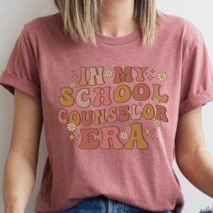 May include: A pink t-shirt with the text "In My School Counselor Era" in a retro font with flowers and stars.
