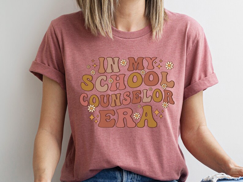 May include: A pink t-shirt with the text "In My School Counselor Era" in a retro font with flowers and stars.