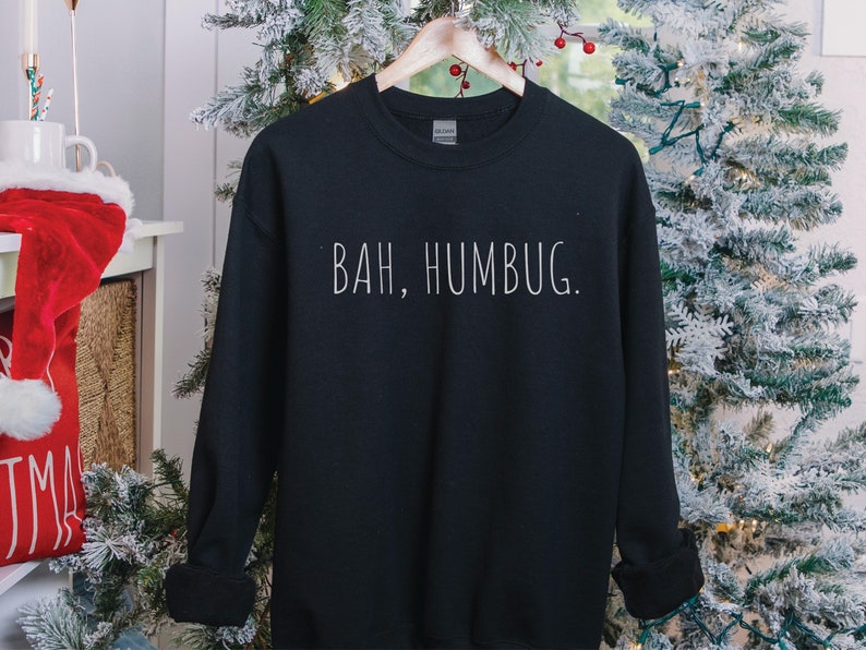 May include: Black sweatshirt with white text that reads "BAH, HUMBUG." The sweatshirt is hanging on a hanger in front of a Christmas tree.