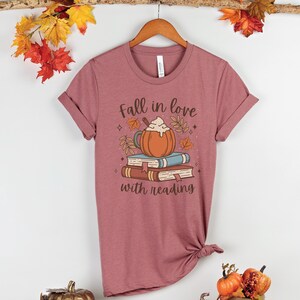 Pode incluir: Uma camiseta malva com um gr&aacute;fico de um latte de especiarias de ab&oacute;bora em cima de uma pilha de livros. O texto "Fall in love with reading" est&aacute; impresso abaixo do gr&aacute;fico.