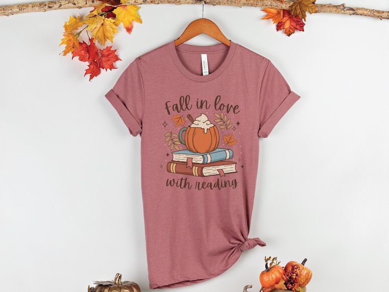 Pode incluir: Uma camiseta malva com um gr&aacute;fico de um latte de especiarias de ab&oacute;bora em cima de uma pilha de livros. O texto "Fall in love with reading" est&aacute; impresso abaixo do gr&aacute;fico.