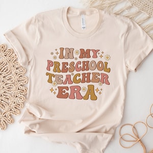 May include: A beige t-shirt with the text "IN MY PRESCHOOL TEACHER ERA" in a retro font with pink, orange, and brown colors. The text is surrounded by small white flowers and gold glitter.