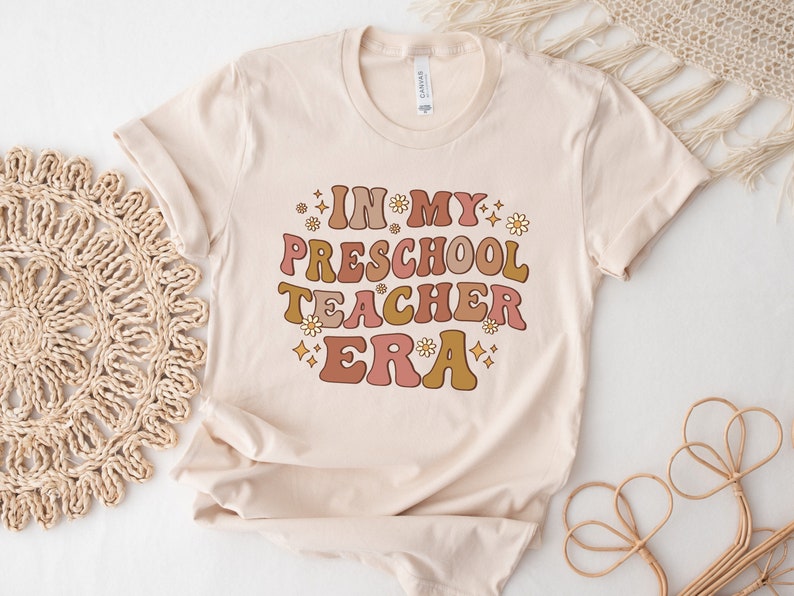 May include: A beige t-shirt with the text "IN MY PRESCHOOL TEACHER ERA" in a retro font with pink, orange, and brown colors. The text is surrounded by small white flowers and gold glitter.