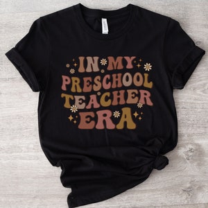May include: Black t-shirt with the text "In My Preschool Teacher Era" in a retro font with brown, pink, and gold accents and small flower graphics.
