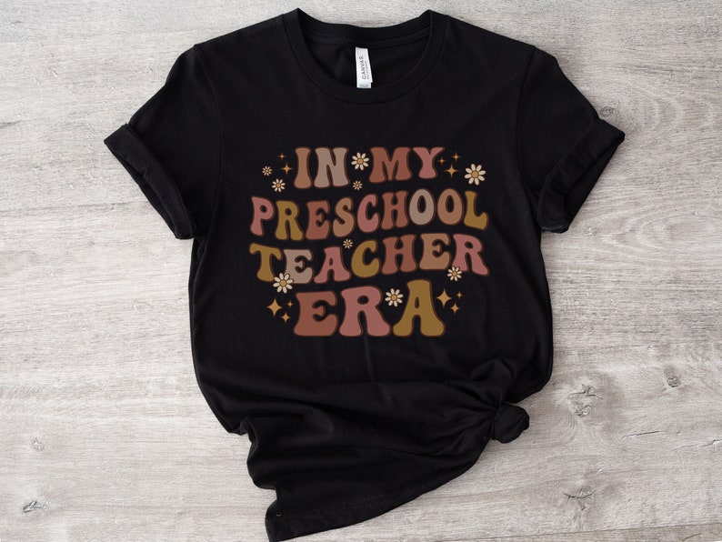 May include: Black t-shirt with the text "In My Preschool Teacher Era" in a retro font with brown, pink, and gold accents and small flower graphics.