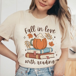 Pode incluir: Uma camiseta bege com o texto "Fall in love with reading" e um gr&aacute;fico de um latte de especiarias de ab&oacute;bora em cima de uma pilha de livros.