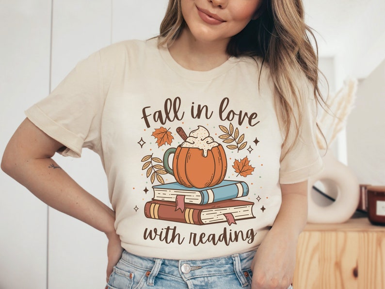Pode incluir: Uma camiseta bege com o texto "Fall in love with reading" e um gr&aacute;fico de um latte de especiarias de ab&oacute;bora em cima de uma pilha de livros.
