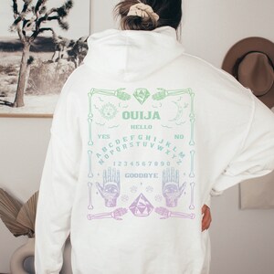 May include: White hoodie with a Ouija board design on the back. The design features a sun, moon, hands, and a planchette, with the words "Ouija", "Hello", "Yes", "No", and "Goodbye". The design is in shades of green and purple.