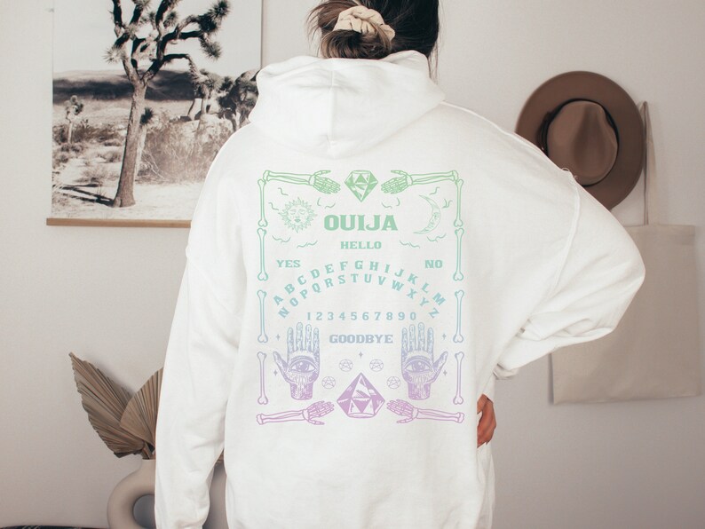 May include: White hoodie with a Ouija board design on the back. The design features a sun, moon, hands, and a planchette, with the words "Ouija", "Hello", "Yes", "No", and "Goodbye". The design is in shades of green and purple.