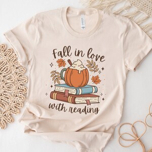 Pode incluir: Uma camiseta bege com um design gr&aacute;fico de um latte de ab&oacute;bora em cima de uma pilha de livros. O texto "Fall in love with reading" est&aacute; impresso abaixo do gr&aacute;fico.