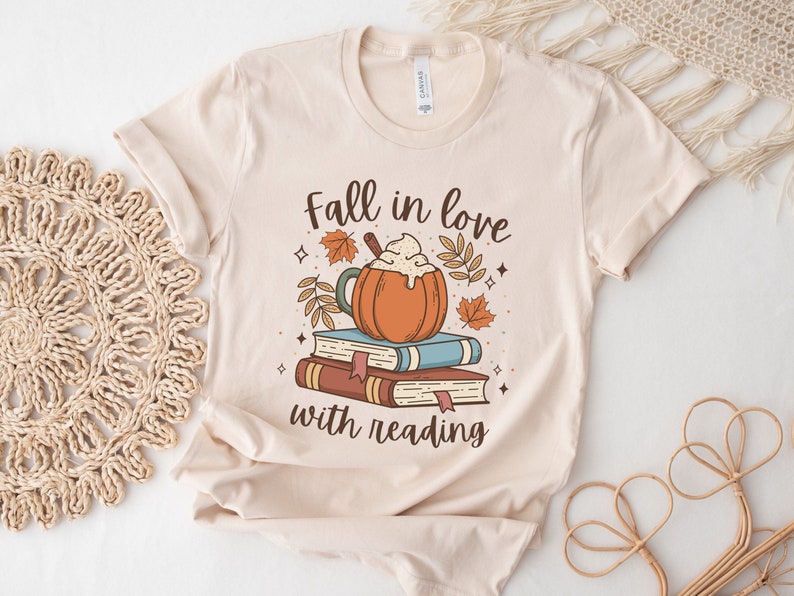 Pode incluir: Uma camiseta bege com um design gr&aacute;fico de um latte de ab&oacute;bora em cima de uma pilha de livros. O texto "Fall in love with reading" est&aacute; impresso abaixo do gr&aacute;fico.