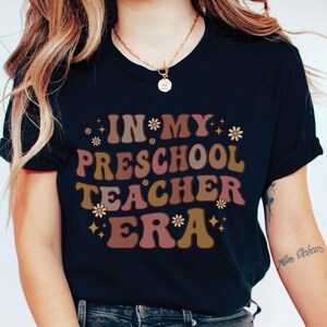 May include: A black t-shirt with the text "IN MY PRESCHOOL TEACHER ERA" in brown, pink, and gold letters with a retro font. The text is surrounded by small white flowers and gold stars.