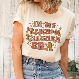 May include: A beige t-shirt with the text "In My Preschool Teacher Era" in a retro font with pink, brown, and gold colors. The text is surrounded by small white flowers and gold glitter.