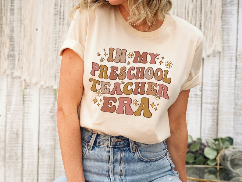 May include: A beige t-shirt with the text "In My Preschool Teacher Era" in a retro font with pink, brown, and gold colors. The text is surrounded by small white flowers and gold glitter.