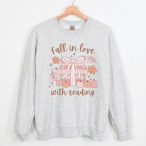 Pu&ograve; includere: Una felpa grigia con un design grafico di una pila di libri legati con un nastro rosa e circondati da foglie autunnali. Il testo "Fall in love with reading" &egrave; stampato sotto il grafico.