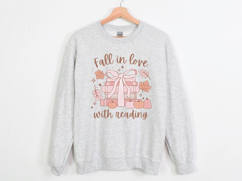 Pu&ograve; includere: Una felpa grigia con un design grafico di una pila di libri legati con un nastro rosa e circondati da foglie autunnali. Il testo "Fall in love with reading" &egrave; stampato sotto il grafico.