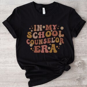 May include: Black t-shirt with the text "In My School Counselor Era" in a retro font with pink, brown, and gold accents and small flower graphics.