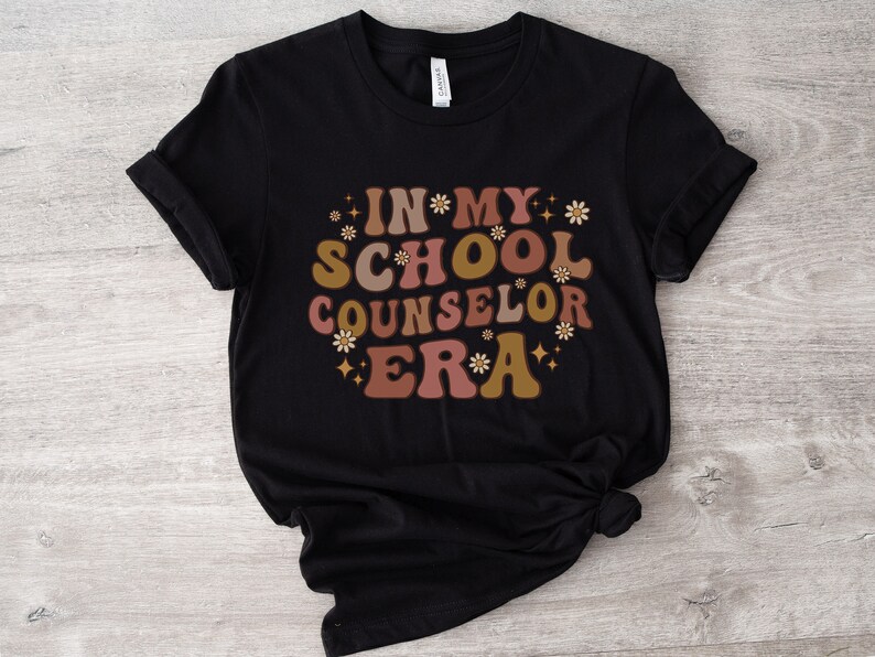 May include: Black t-shirt with the text "In My School Counselor Era" in a retro font with pink, brown, and gold accents and small flower graphics.