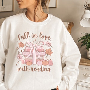 Pu&ograve; includere: Felpa bianca con un design grafico rosa e marrone che dice "Innamorati della lettura". La grafica presenta una pila di libri con un fiocco rosa, foglie autunnali e zucche.