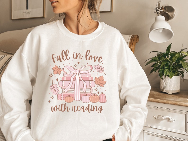 Pu&ograve; includere: Felpa bianca con un design grafico rosa e marrone che dice "Innamorati della lettura". La grafica presenta una pila di libri con un fiocco rosa, foglie autunnali e zucche.