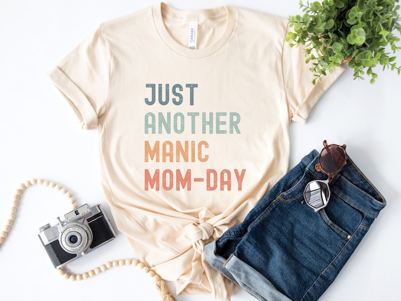 May include: A beige t-shirt with the text "JUST ANOTHER MANIC MOM-DAY" in a vintage style font with colourful accents.