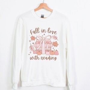 Pu&ograve; includere: Felpa bianca con una grafica di una pila di libri legati con un nastro rosa e circondati da foglie autunnali e zucche. Il testo "Fall in love with reading" &egrave; stampato sotto la grafica.
