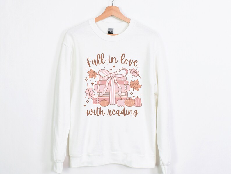Pu&ograve; includere: Felpa bianca con una grafica di una pila di libri legati con un nastro rosa e circondati da foglie autunnali e zucche. Il testo "Fall in love with reading" &egrave; stampato sotto la grafica.