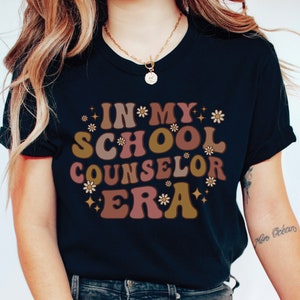 May include: A black t-shirt with the text "In My School Counselor Era" in a retro font with brown, pink, and gold colors and white flowers.