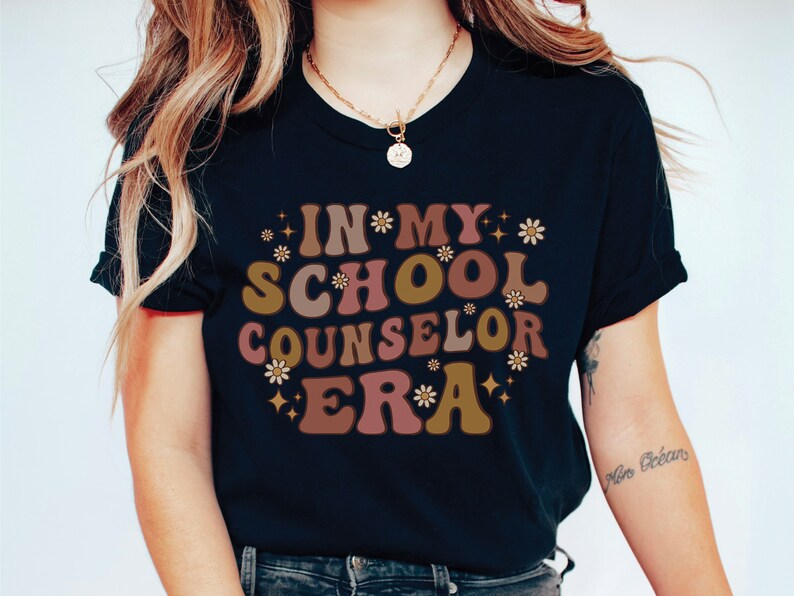 May include: A black t-shirt with the text "In My School Counselor Era" in a retro font with brown, pink, and gold colors and white flowers.