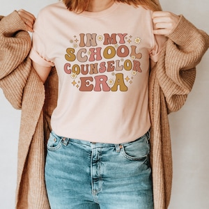 May include: A pink t-shirt with the text "In My School Counselor Era" in a retro font with flowers and stars. The shirt is being worn by a person with a brown cardigan.
