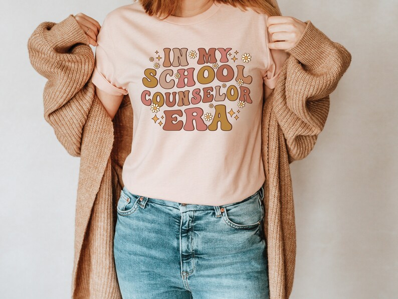 May include: A pink t-shirt with the text "In My School Counselor Era" in a retro font with flowers and stars. The shirt is being worn by a person with a brown cardigan.