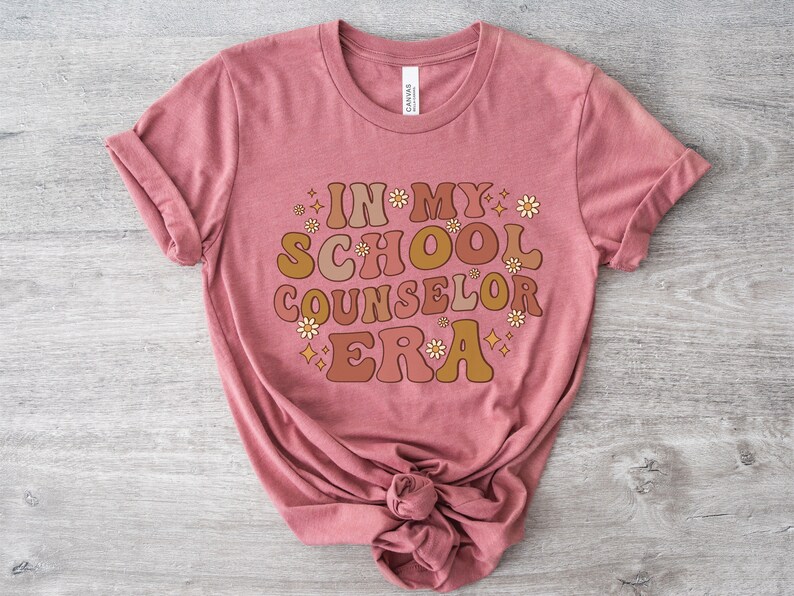 May include: A pink t-shirt with the text "In My School Counselor Era" in a retro font with flowers and sparkles.