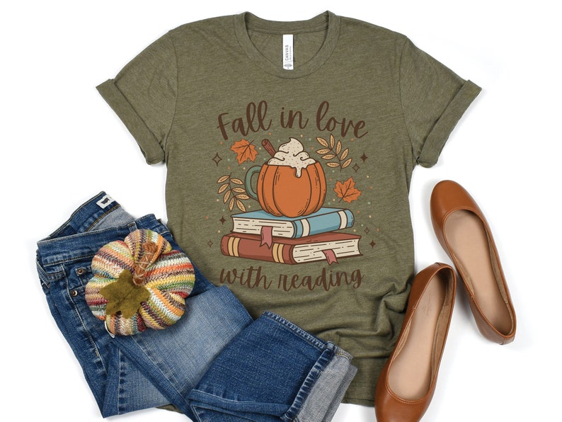 Pode incluir: Uma camiseta verde com o texto "Fall in love with reading" e uma imagem de desenho animado de um latte de especiarias de ab&oacute;bora com chantilly em cima de uma pilha de livros.