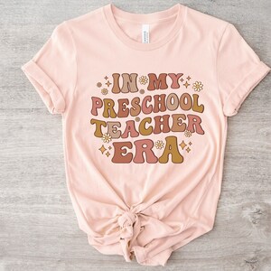 May include: A pink t-shirt with the text "IN MY PRESCHOOL TEACHER ERA" in brown, orange, and pink letters with a retro font. The text is surrounded by small white flowers and gold stars.