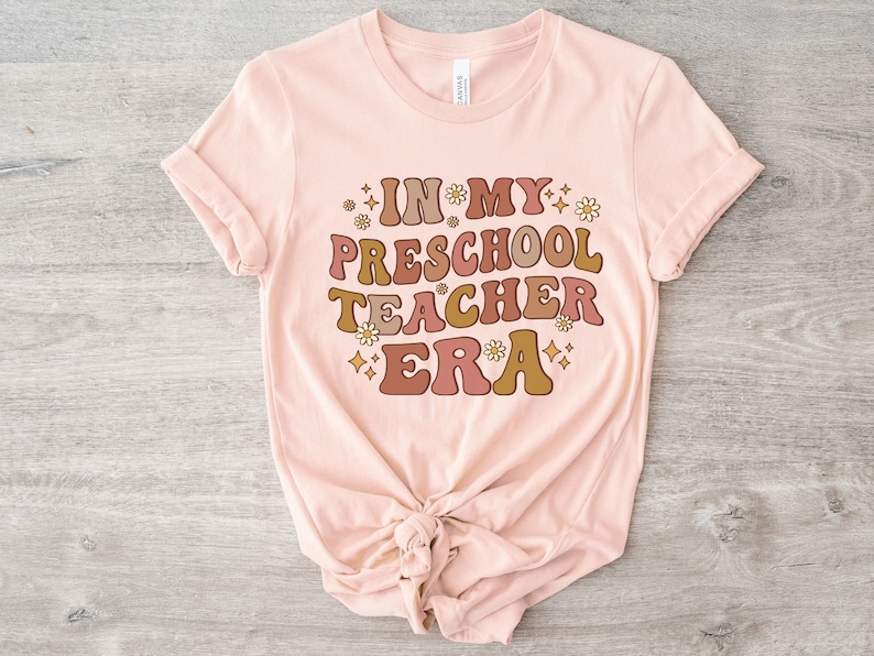 May include: A pink t-shirt with the text "IN MY PRESCHOOL TEACHER ERA" in brown, orange, and pink letters with a retro font. The text is surrounded by small white flowers and gold stars.