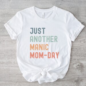 May include: White t-shirt with the text "JUST ANOTHER MANIC MOM-DAY" in a rainbow gradient.
