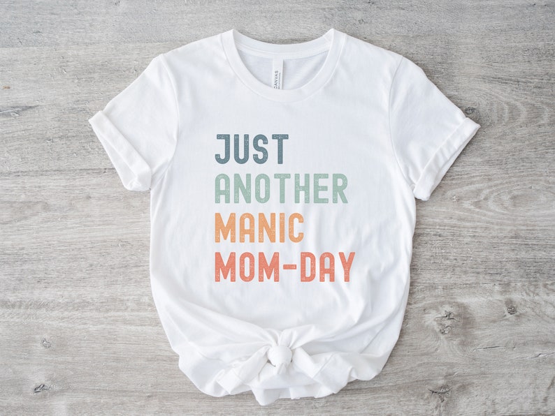 May include: White t-shirt with the text "JUST ANOTHER MANIC MOM-DAY" in a rainbow gradient.