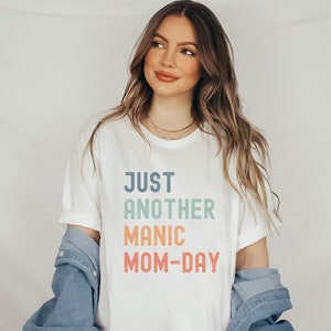May include: White t-shirt with the text "JUST ANOTHER MANIC MOM-DAY" in a rainbow gradient.