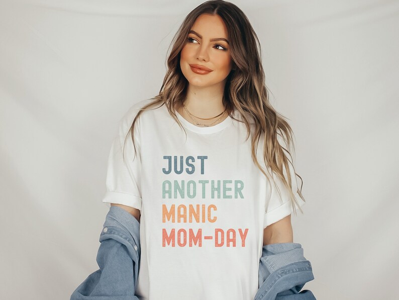 May include: White t-shirt with the text "JUST ANOTHER MANIC MOM-DAY" in a rainbow gradient.