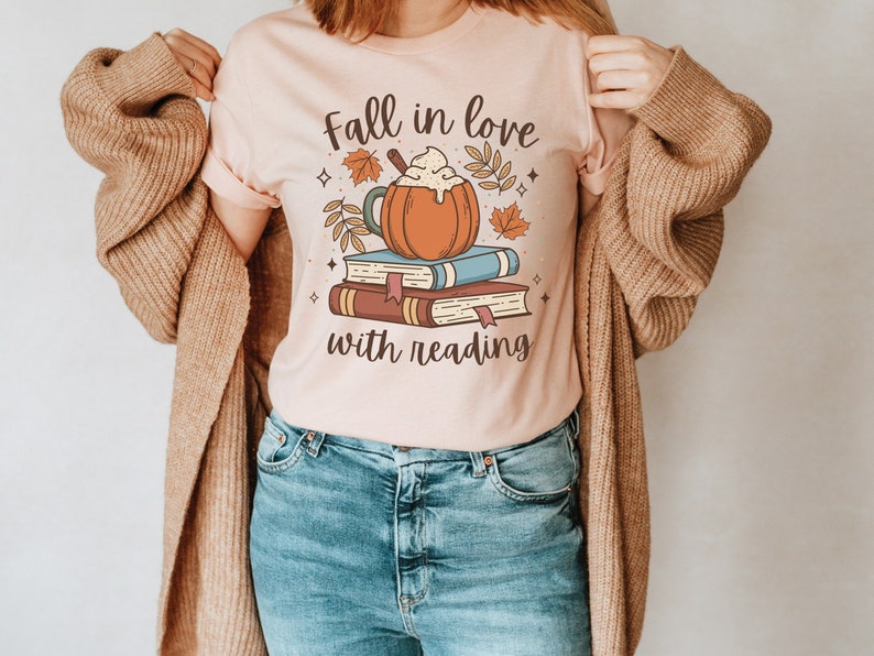 Pode incluir: Uma camiseta rosa com o texto "Fall in love with reading" e uma ilustra&ccedil;&atilde;o de desenho animado de um latte de especiarias de ab&oacute;bora em cima de uma pilha de livros.